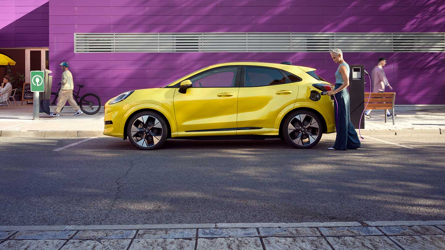 Seitenansicht eines parkenden gelben elektrischen Ford Puma Gen-E®, der an eine öffentliche Ladestation angeschlossen ist.