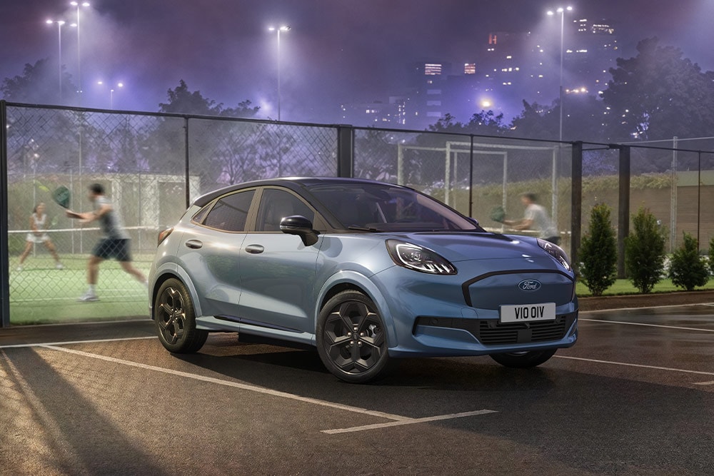 Ein blauer Ford Puma Gen-E parkt neben einem Tennisplatz mit Stadtlichtern im Hintergrund.