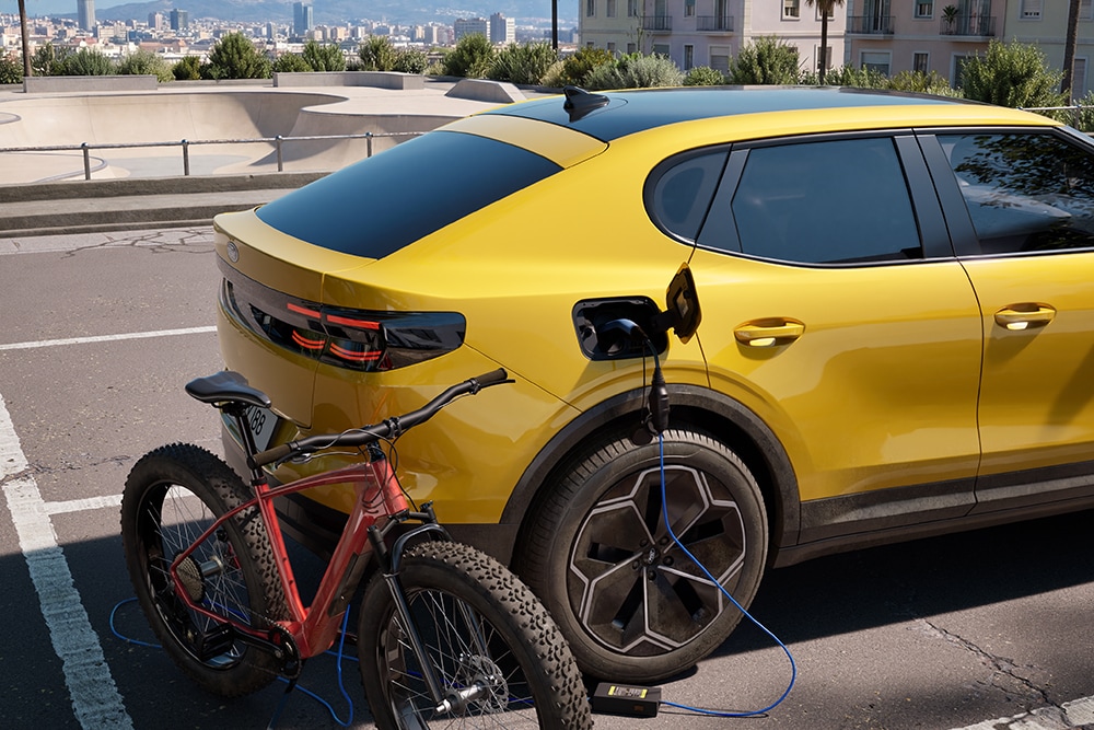 Gelber Ford Capri® parkt und lädt ein rotes Elektrofahrrad in einer Stadtlandschaft