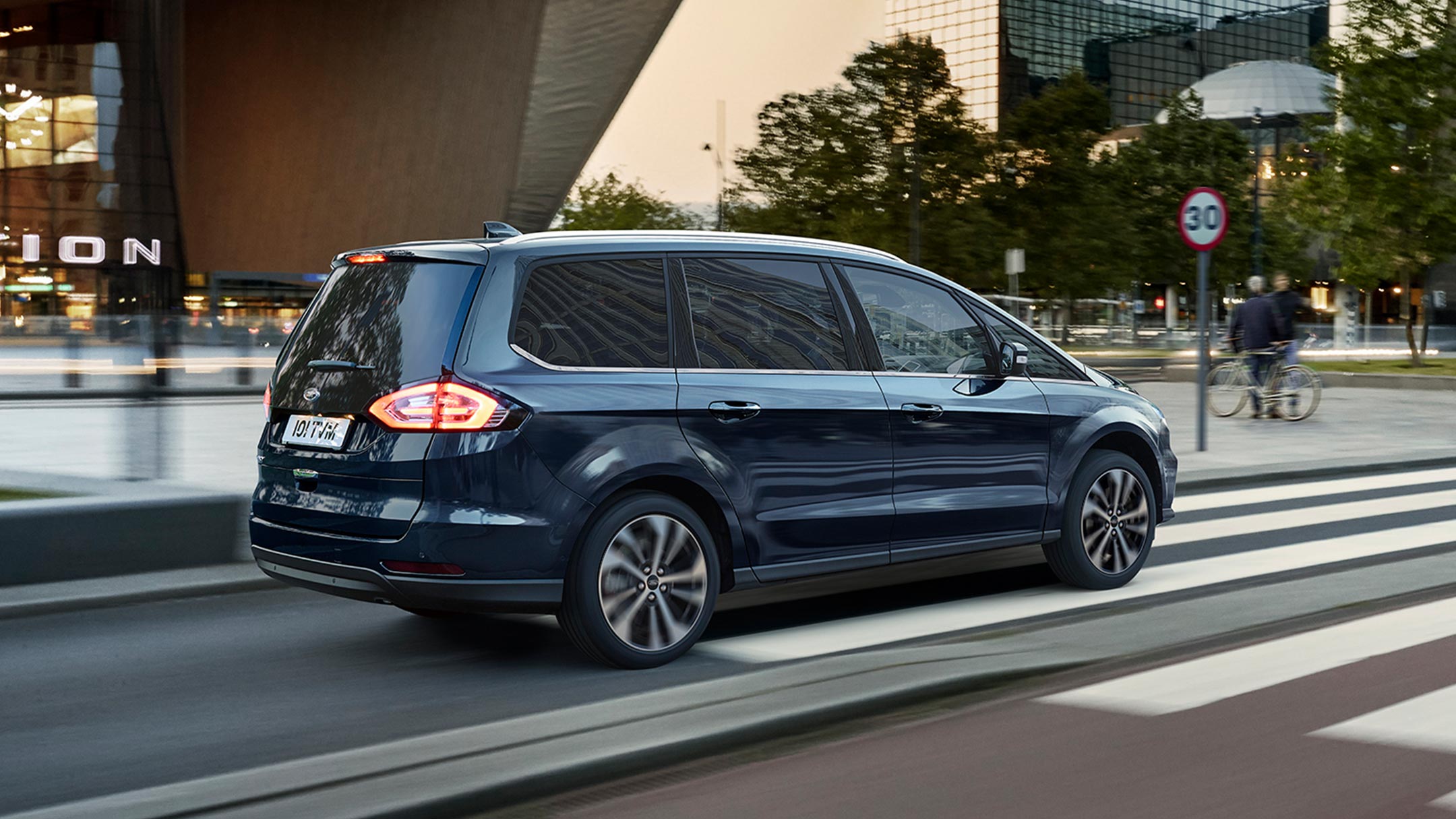 Ford Galaxy Hybrid in Blau. Dreiviertelansicht, fahrend in Stadt über Zebrastreifen vor Bürogebäuden