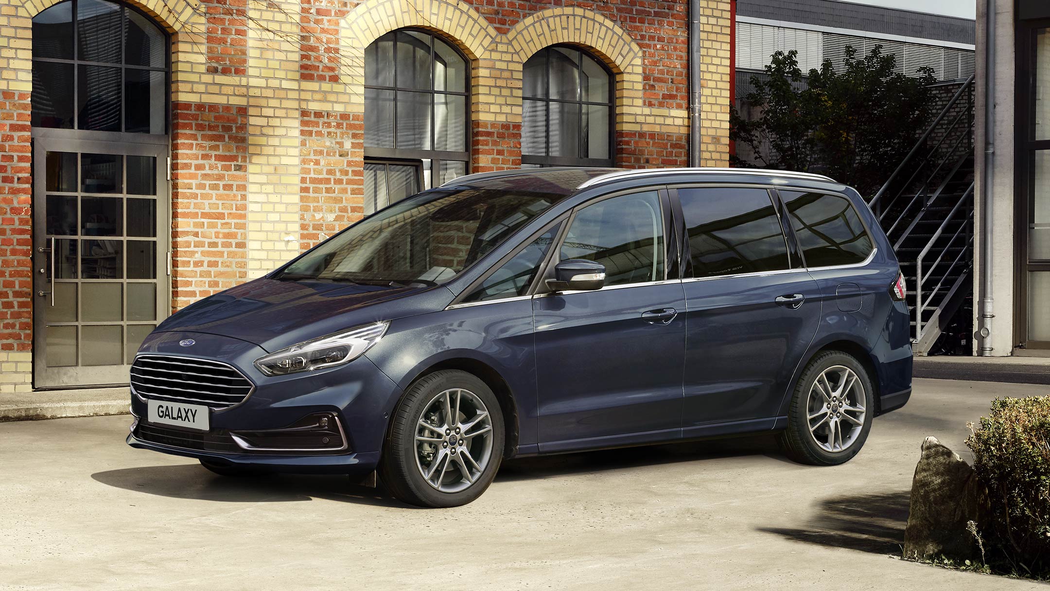 Ford Galaxy Hybrid in Blau. ¾-Frontansicht. Vor einem Ziegelgebäude parkend