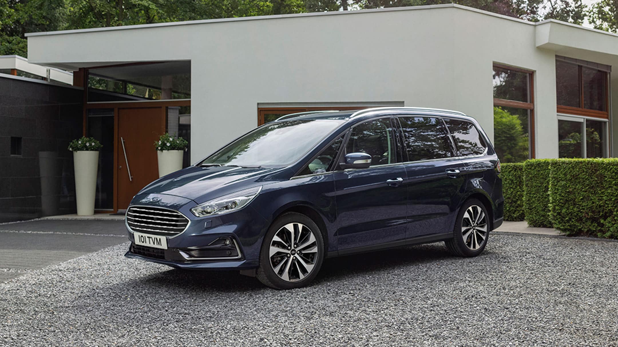 Ford Galaxy Hybrid in Dunkelblau in der 3/4 Frontansicht parkt vor einem Familienhaus