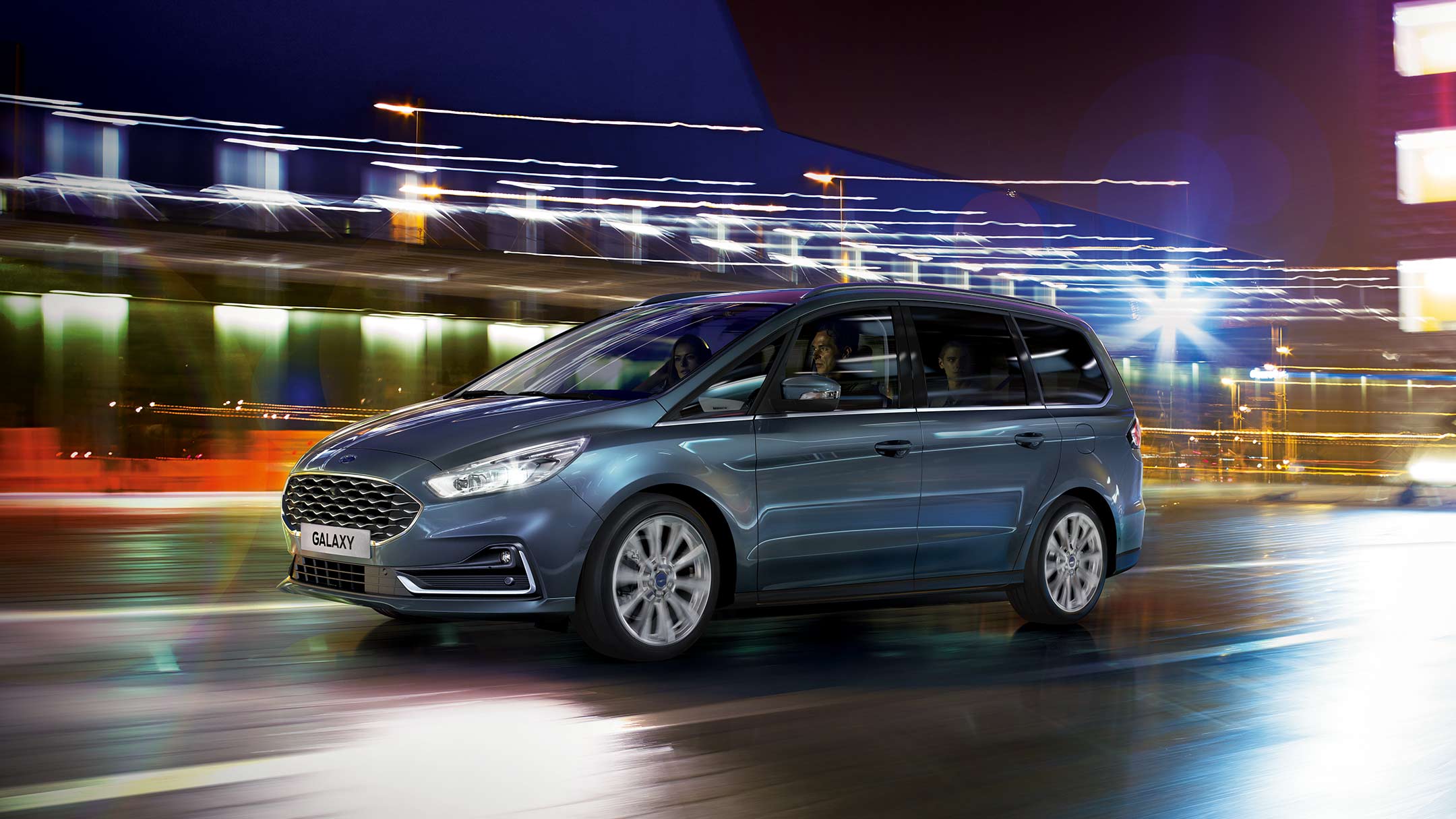 Ford Galaxy Hybrid in Blau. Dreiviertelansicht, fahrend durch eine Stadt bei Nacht