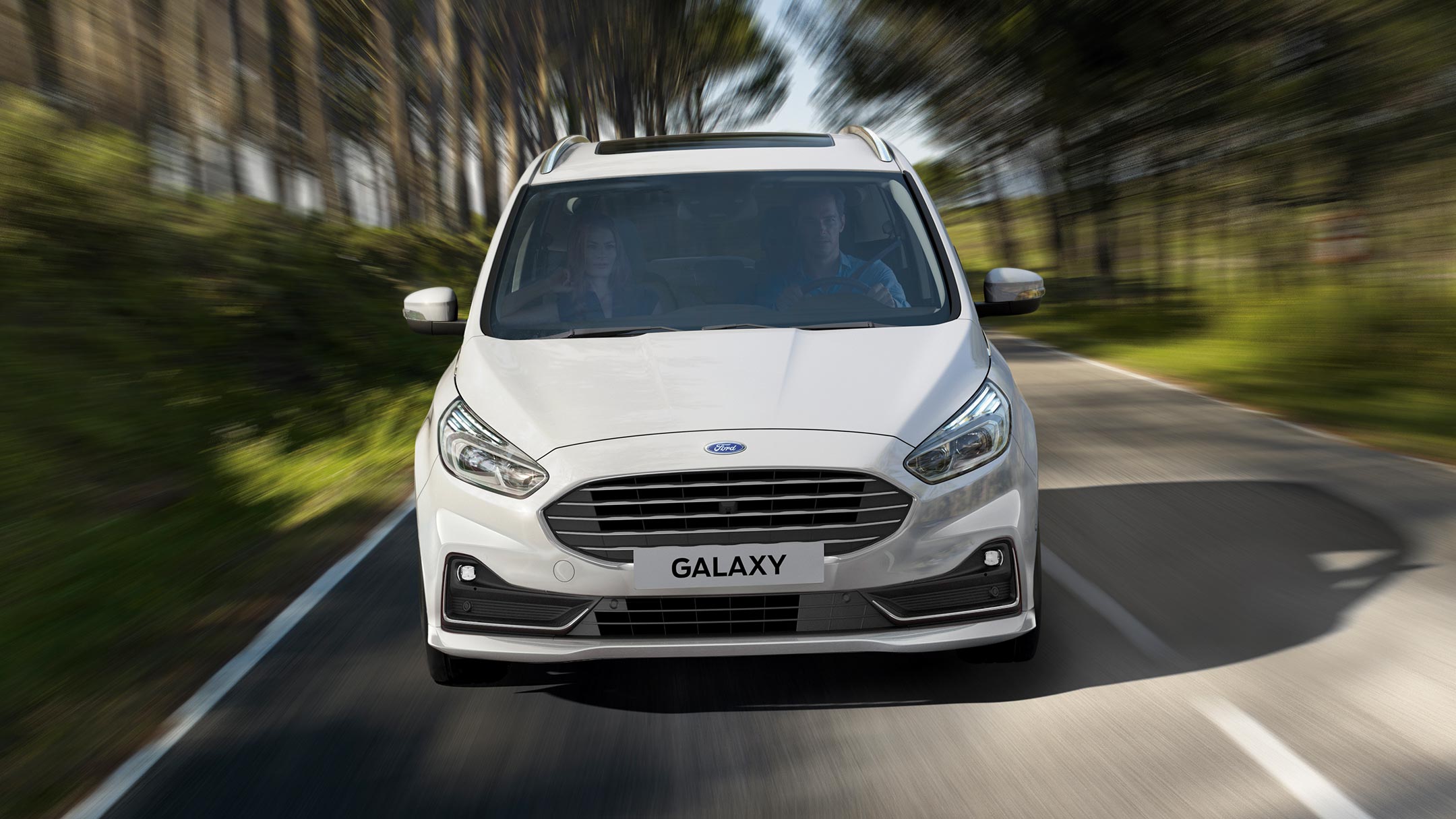 Ford Galaxy Hybrid in Weiß. Frontansicht, fahrend auf Landstraße durch Wald