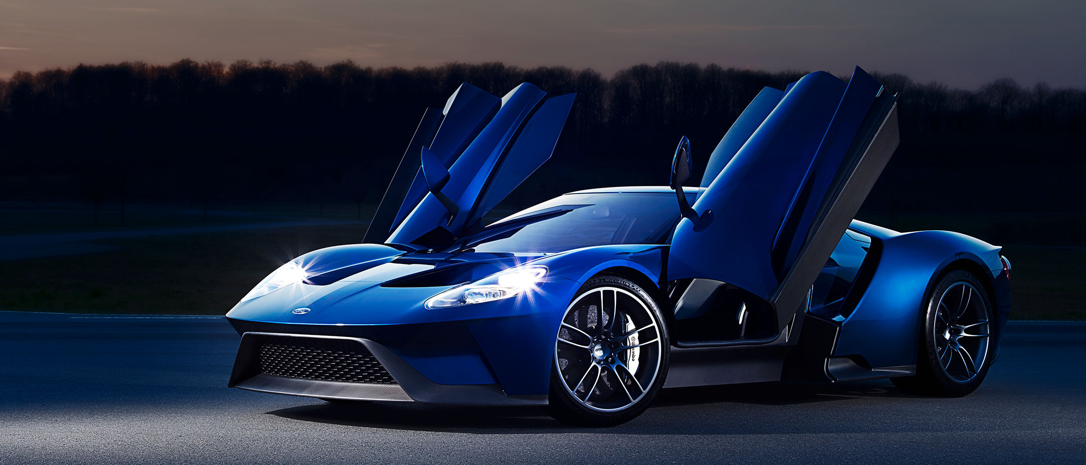 Ford GT Blau Türen geöffnet Scheinwerfer an parkt vor einer Lichterkulisse Ford GT Blau Türen geöffnet Scheinwerfer an parkt vor einer Lichterkulisse