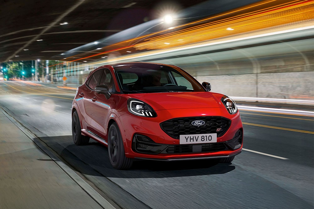 Ein roter, sportlicher Ford Puma ST SUV fährt nachts auf einer hell erleuchteten Stadtstraße.