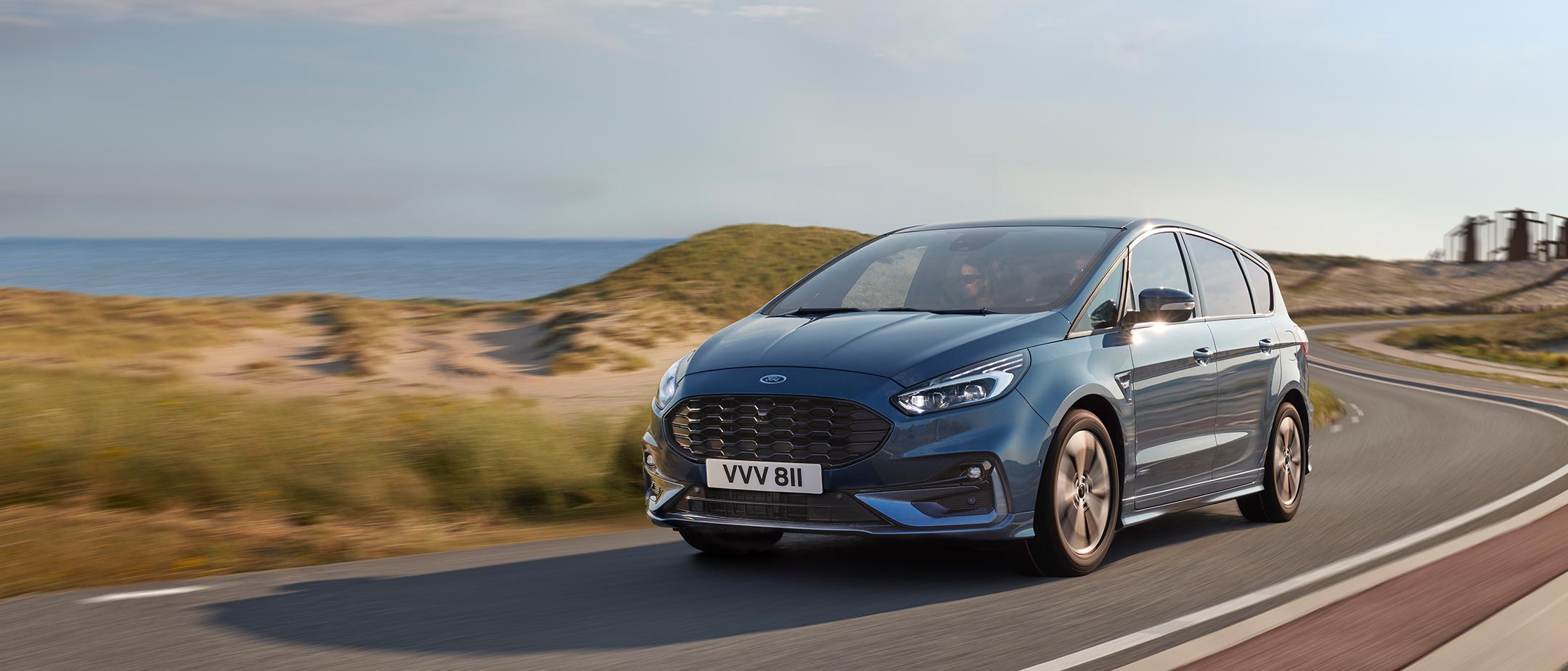 Ford S-MAX Hybrid in Blau. Dreiviertelansicht, fahrend auf Straße in Dünenlandschaft am Meer