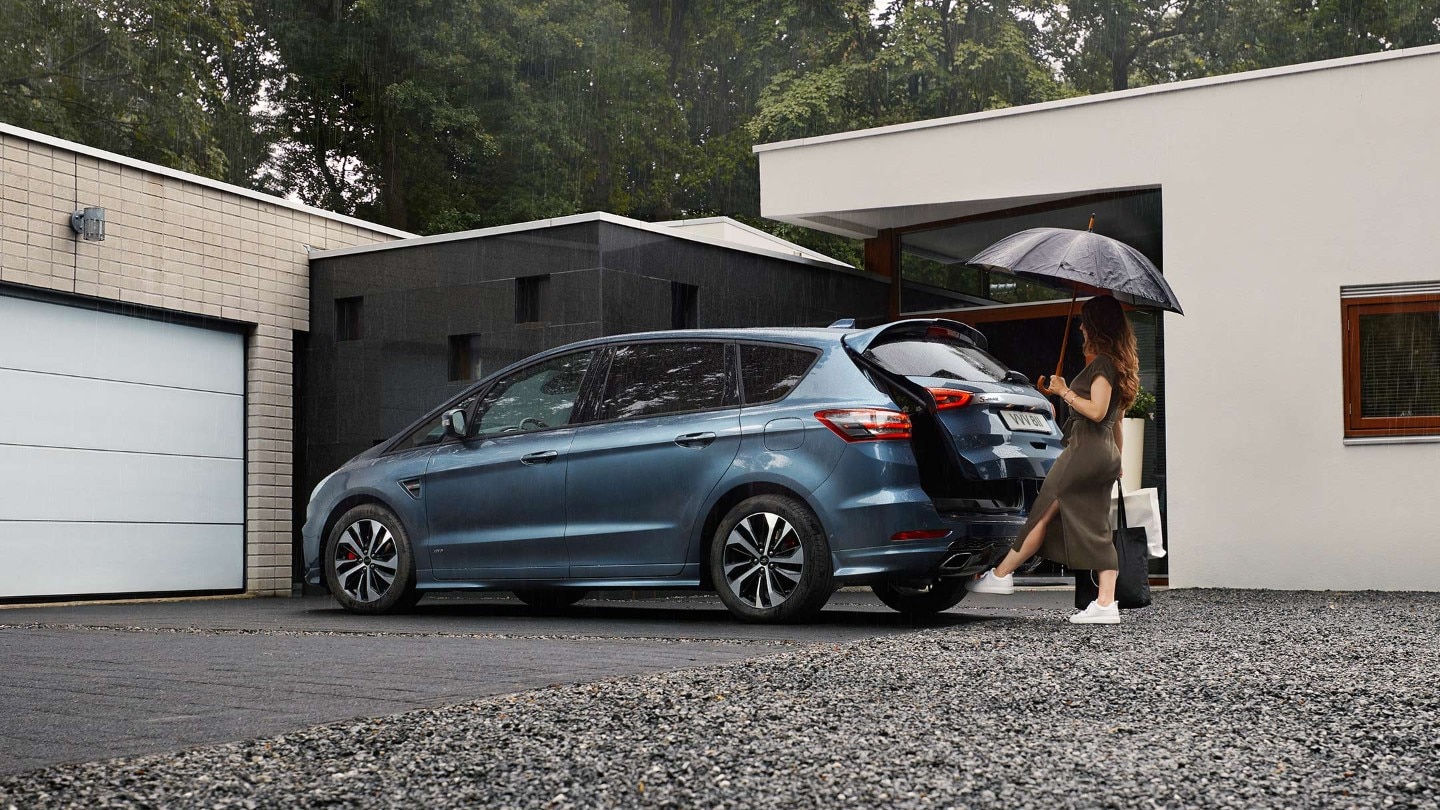 Ford S-MAX Hybrid in Blau. Dreiviertelansicht, stehend vor modernem Haus mit Frau die den Gepäckraum mit Hilfe des Näherungssensors öffnet