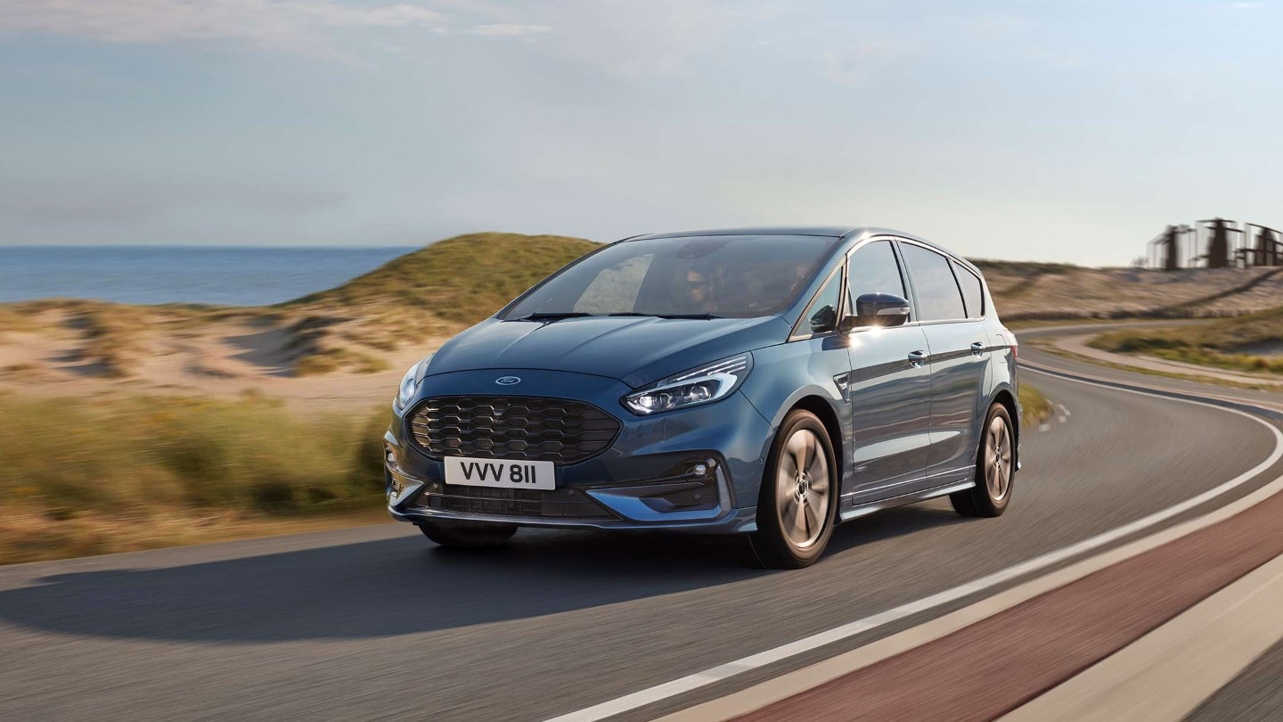 Ford S-MAX Hybrid in Blau. Dreiviertelansicht, fahrend auf Straße in Dünenlandschaft am Meer