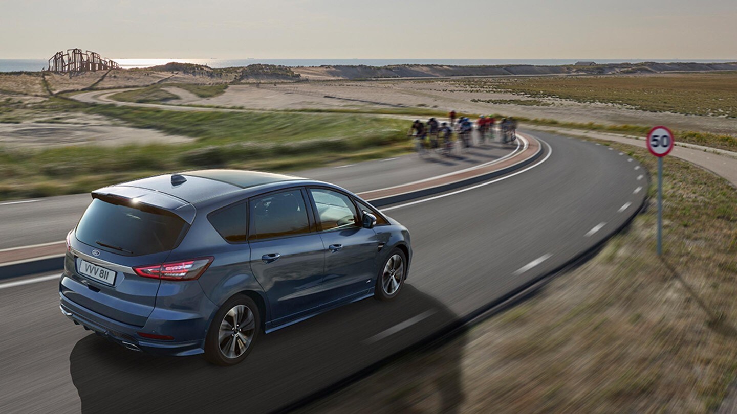Ford S-MAX Hybrid in Blau. Dreiviertelansicht, fahrend über Straße in Dünenlandschaft