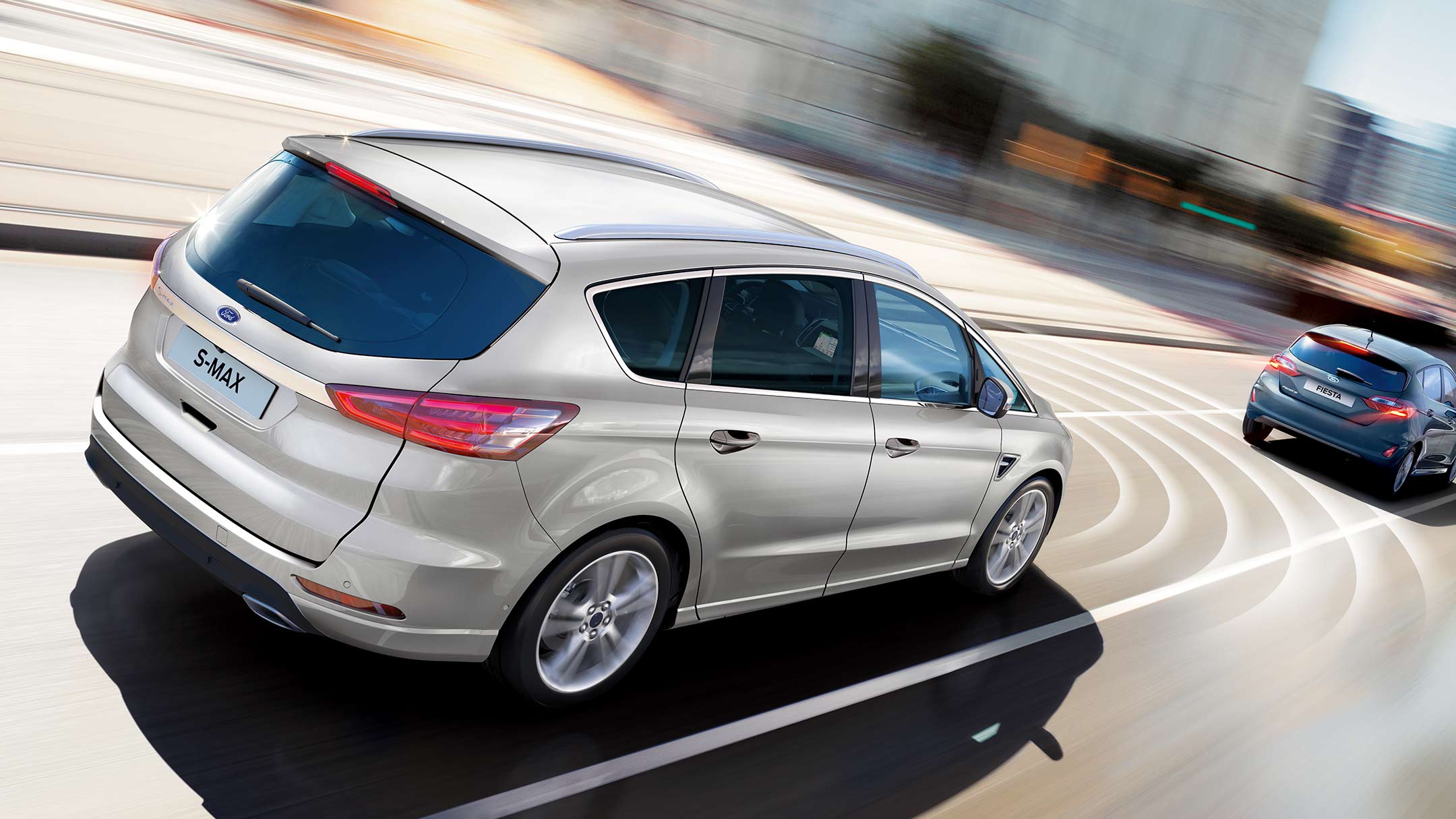 Ford S-MAX Hybrid in Silber. Dreiviertelansicht, fahrend auf Straße hinter blauem Fahrzeug