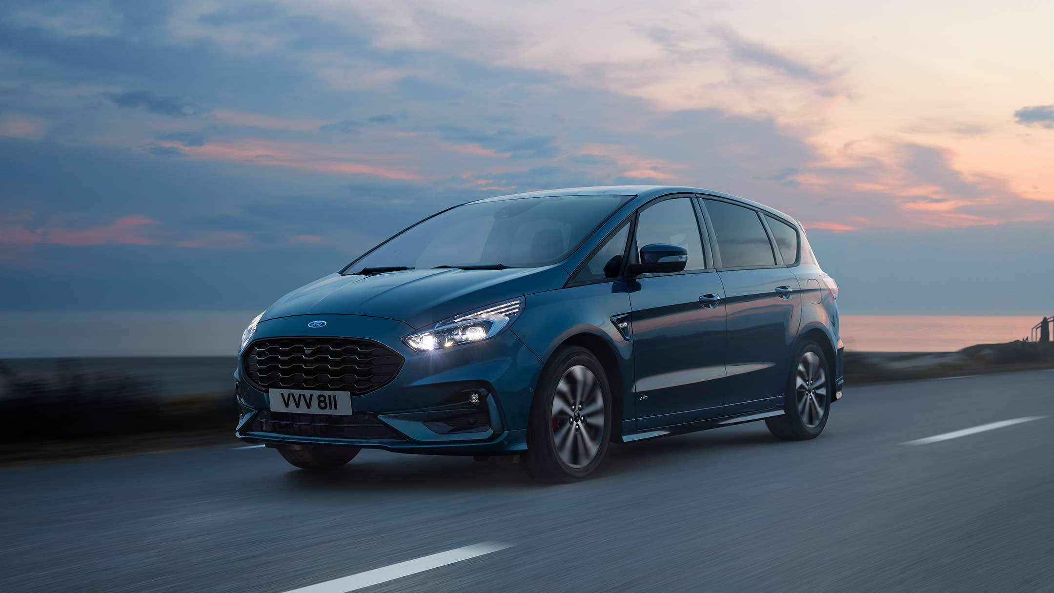 Ford S-MAX Hybrid in Blau. Dreiviertelansicht, fahrend auf Straße am Meer bei Sonnenuntergang
