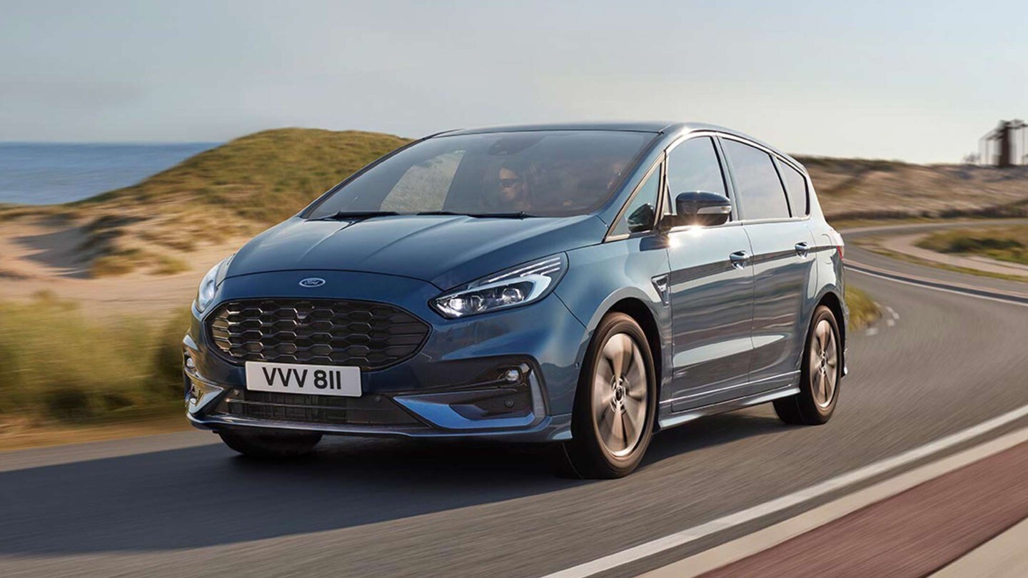 Ford S-MAX Hybrid in Blau. Dreiviertelansicht, fahrend auf Straße in Dünenlandschaft am Meer