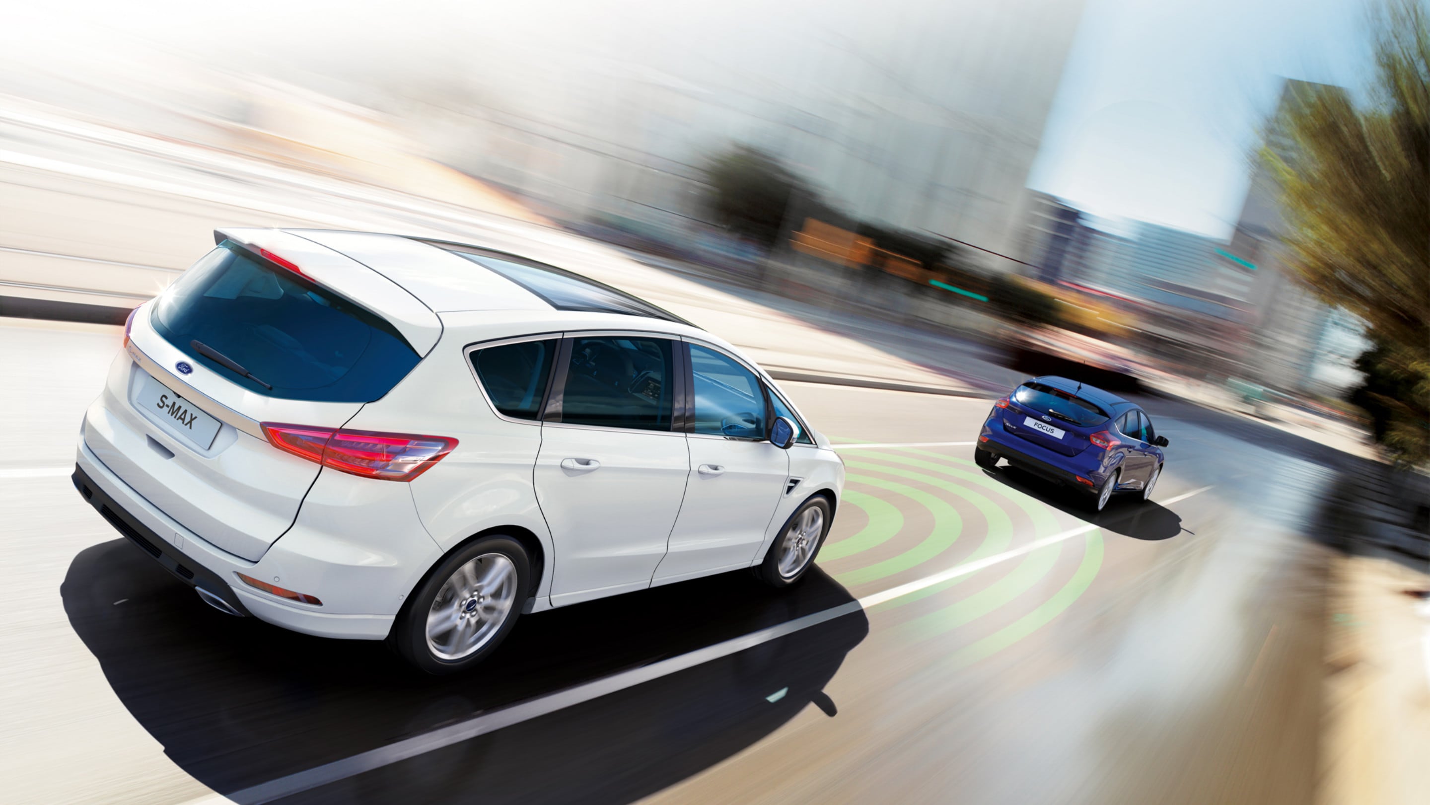 Ford S-MAX Hybrid in Silber. Dreiviertelansicht, fahrend auf Straße hinter blauem Fahrzeug
