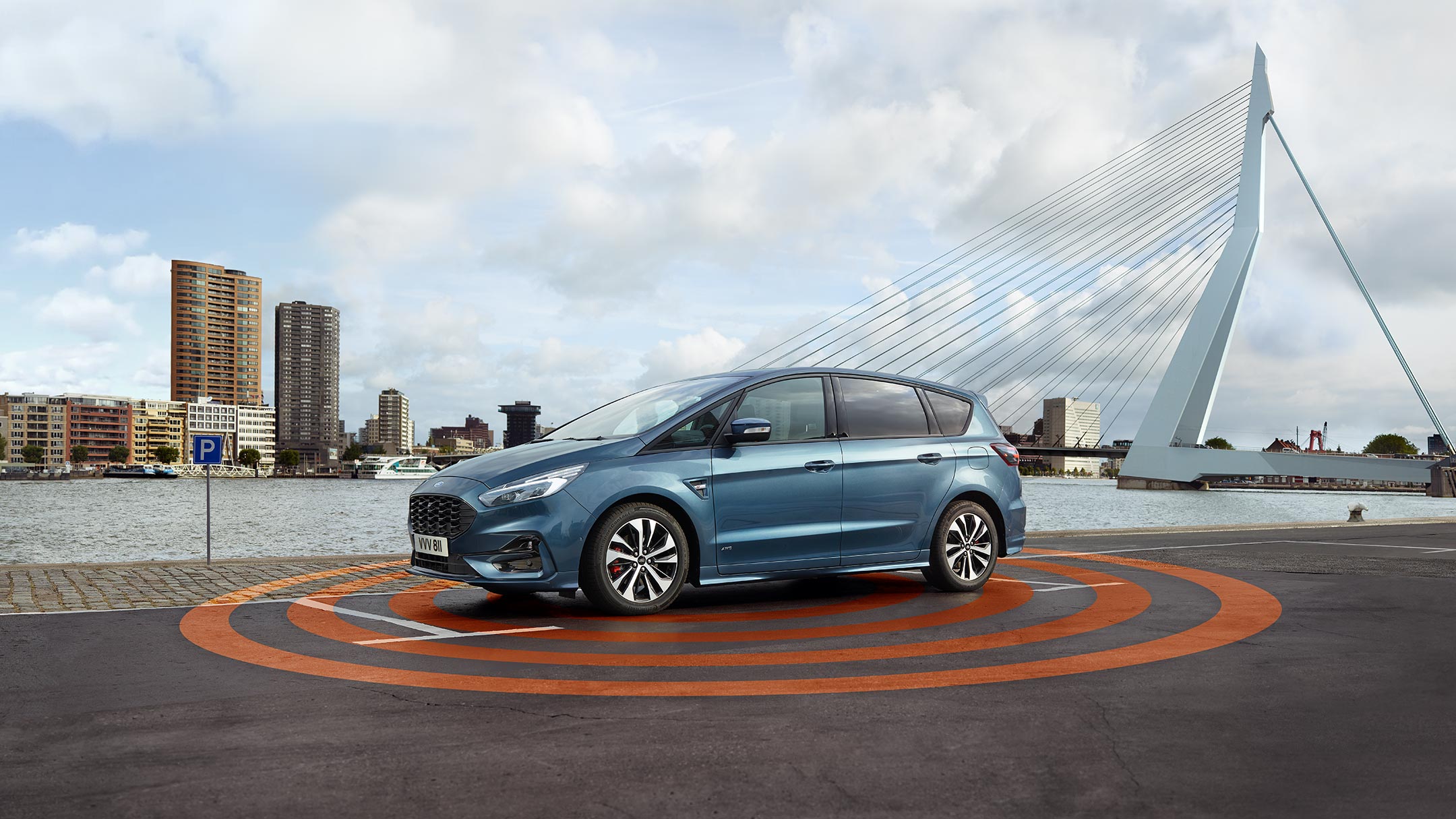 Ford S-MAX Hybrid in Blau. Seitenansicht, stehend auf Parkplatz an Pier in Hafen. Alarmanlage visualisiert mit Kreisen um Fahrzeug
