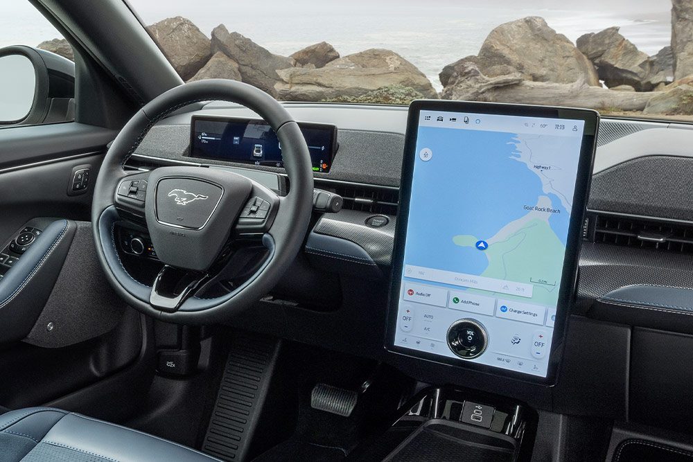 Innenraum eines Ford Mustang Mach-E, zu sehen sind das Armaturenbrett, das Lenkrad und der große Touchscreen mit einer Karte, geparkt an einer felsigen Küste.
