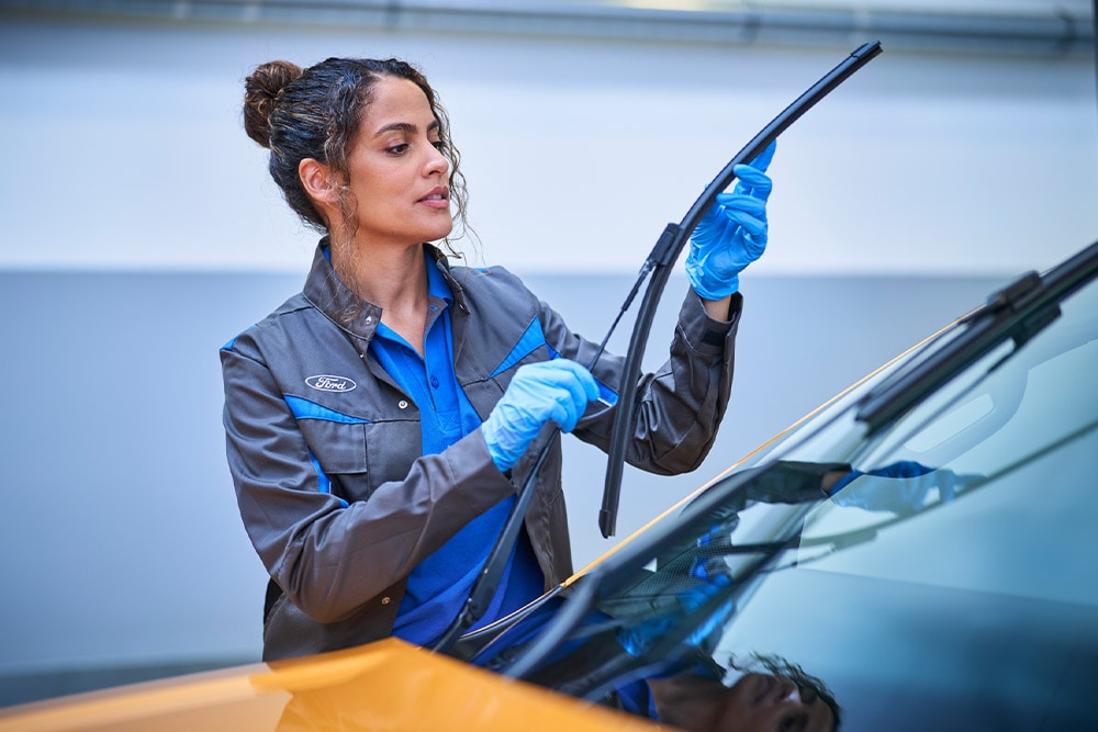 A Ford technician in blue gloves replaces a wiper blade on a car windshield in a service garage.