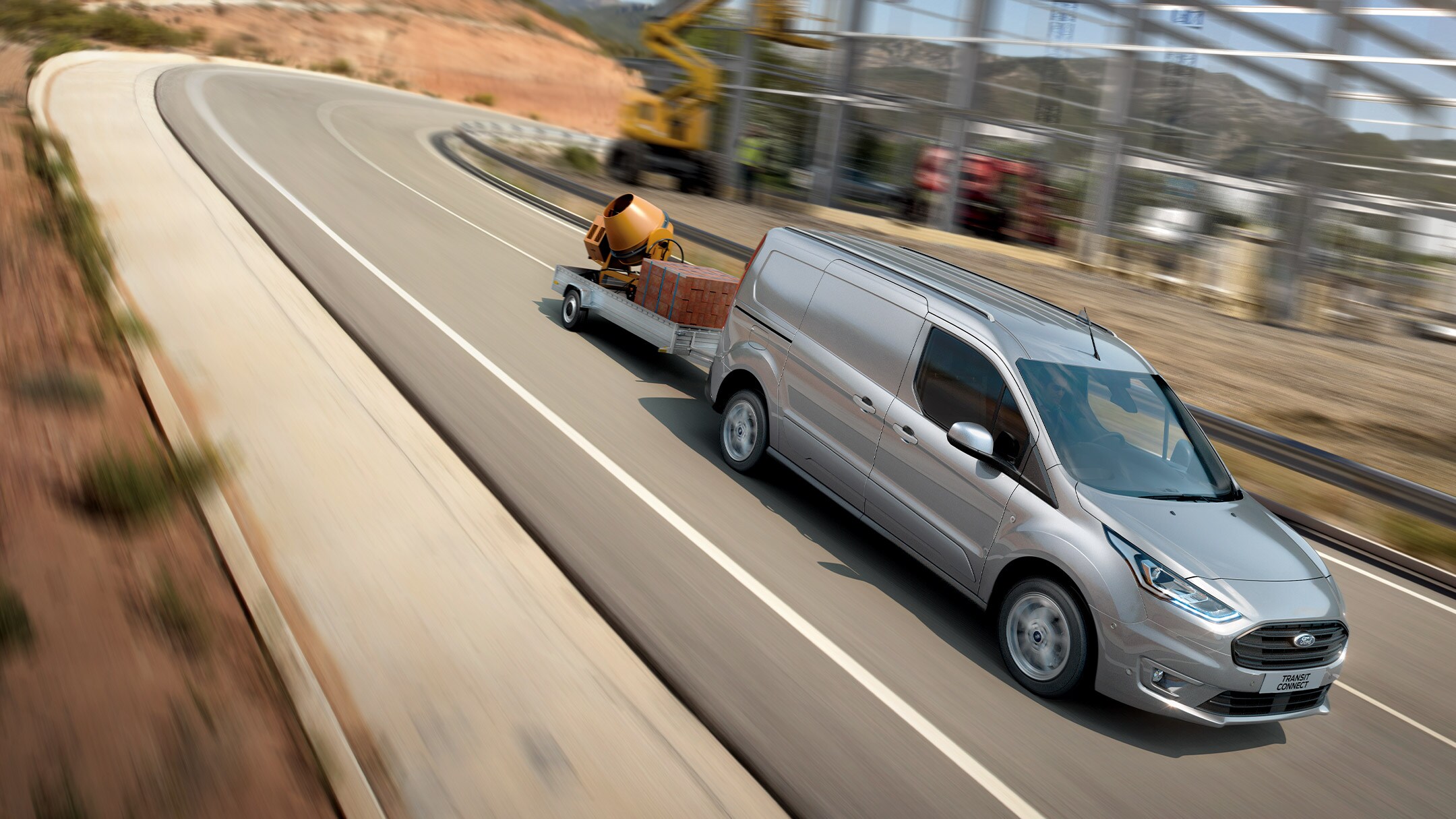 Ford Transit Connect in Silber und Seitenansicht transportiert einen Anhänger bergab auf einer Landstraße