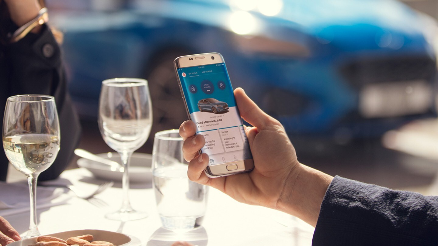 Ford S-MAX Hybrid. Detailansicht Smartphone mit geöffneter FordPass App in der Hand eines Mannes. Im Hintergrund ein blauer Ford Focus