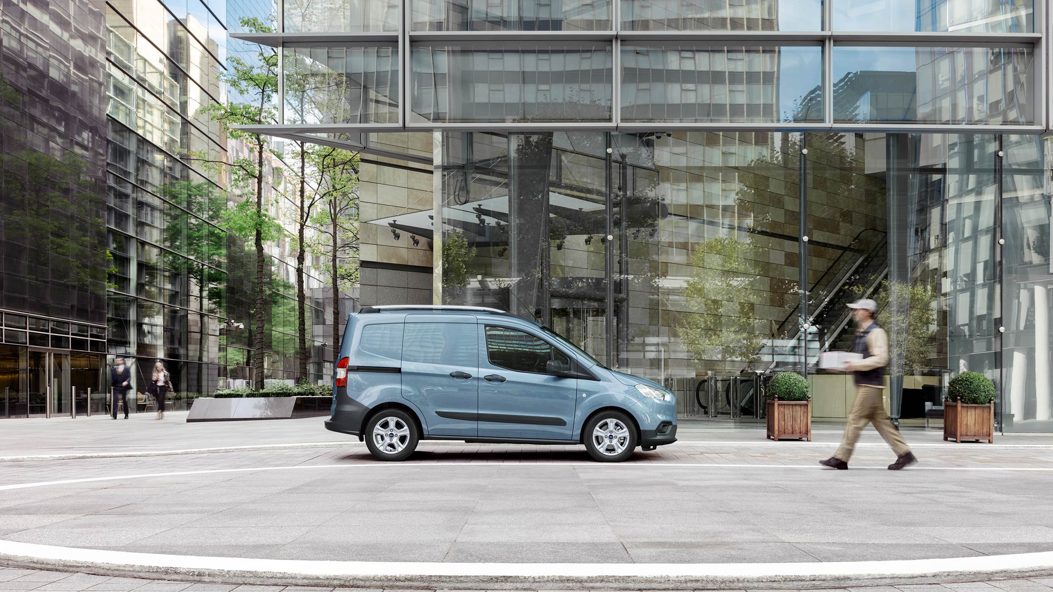 Ford Transit Courier in Grau Seitenansicht parkt vor modernem Gebäude mit Glasfassade