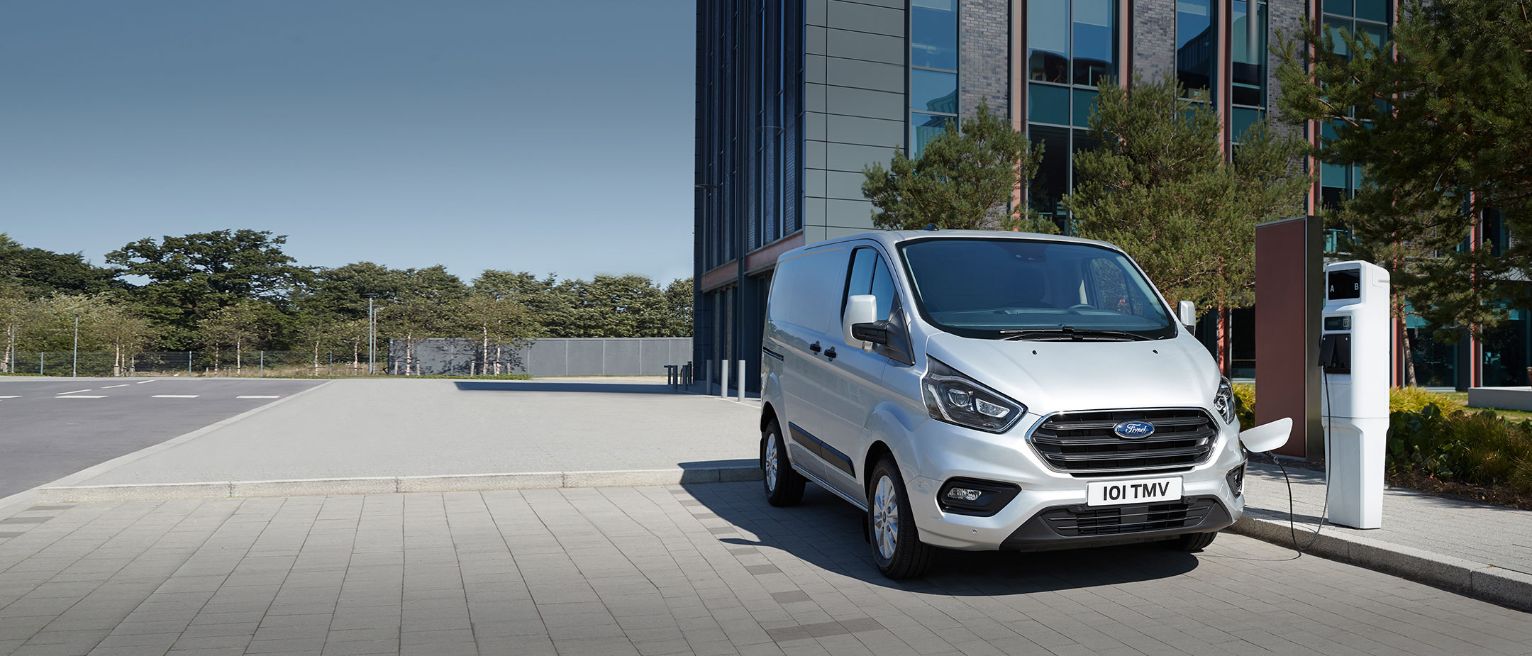 Ford Transit Custom Plug-In Hybrid Silber ¾-Frontansicht parkt an Ladestation Ford Transit Custom Plug-In Hybrid Silber ¾-Frontansicht parkt an Ladestation