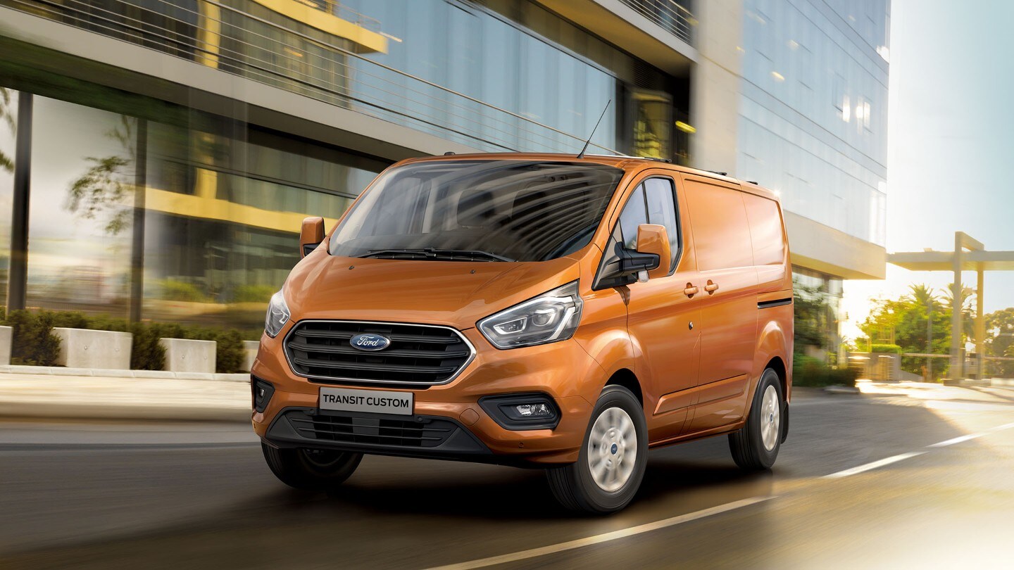 Ford Transit Custom in Orange ¾-Frontansicht in Bewegung vor Gebäude mit Glasfassade