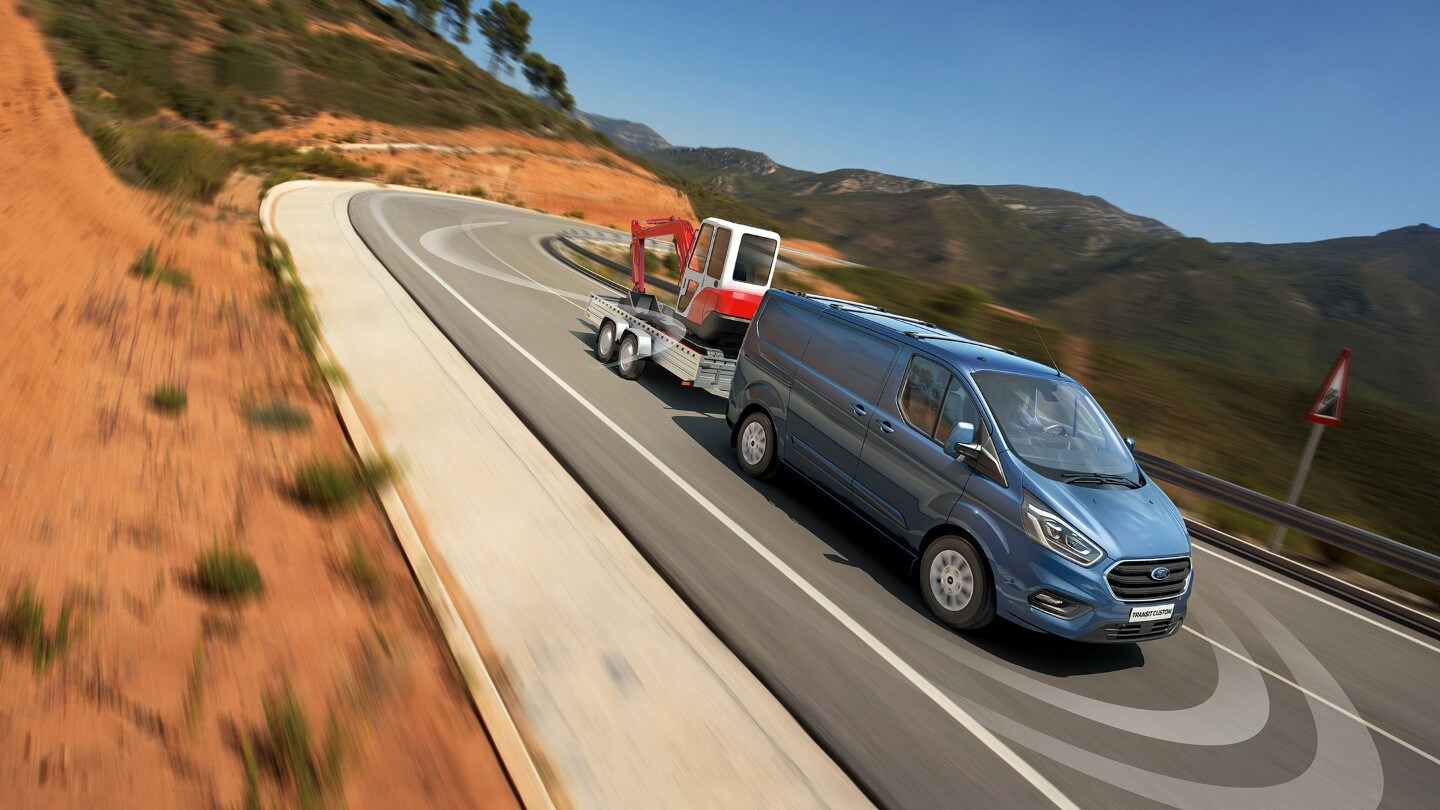 Ford Transit Custom in Blau ¾-Frontansicht mit Anhänger fährt bergab auf Landstraße