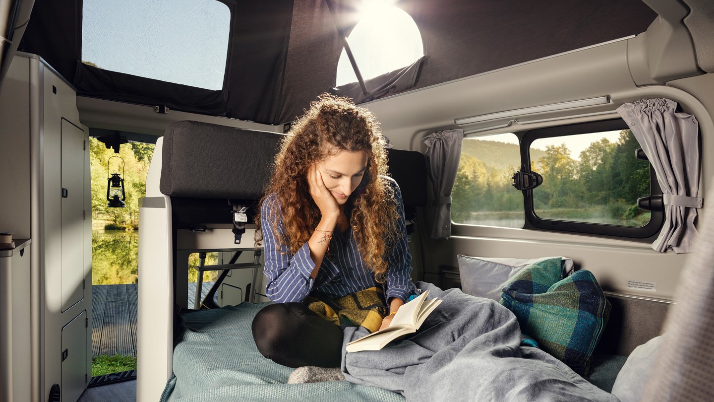 Ford Transit Custom Nugget Aufstelldach Innenraumansicht Frau sitzt auf Doppelbett in Wohnbereich