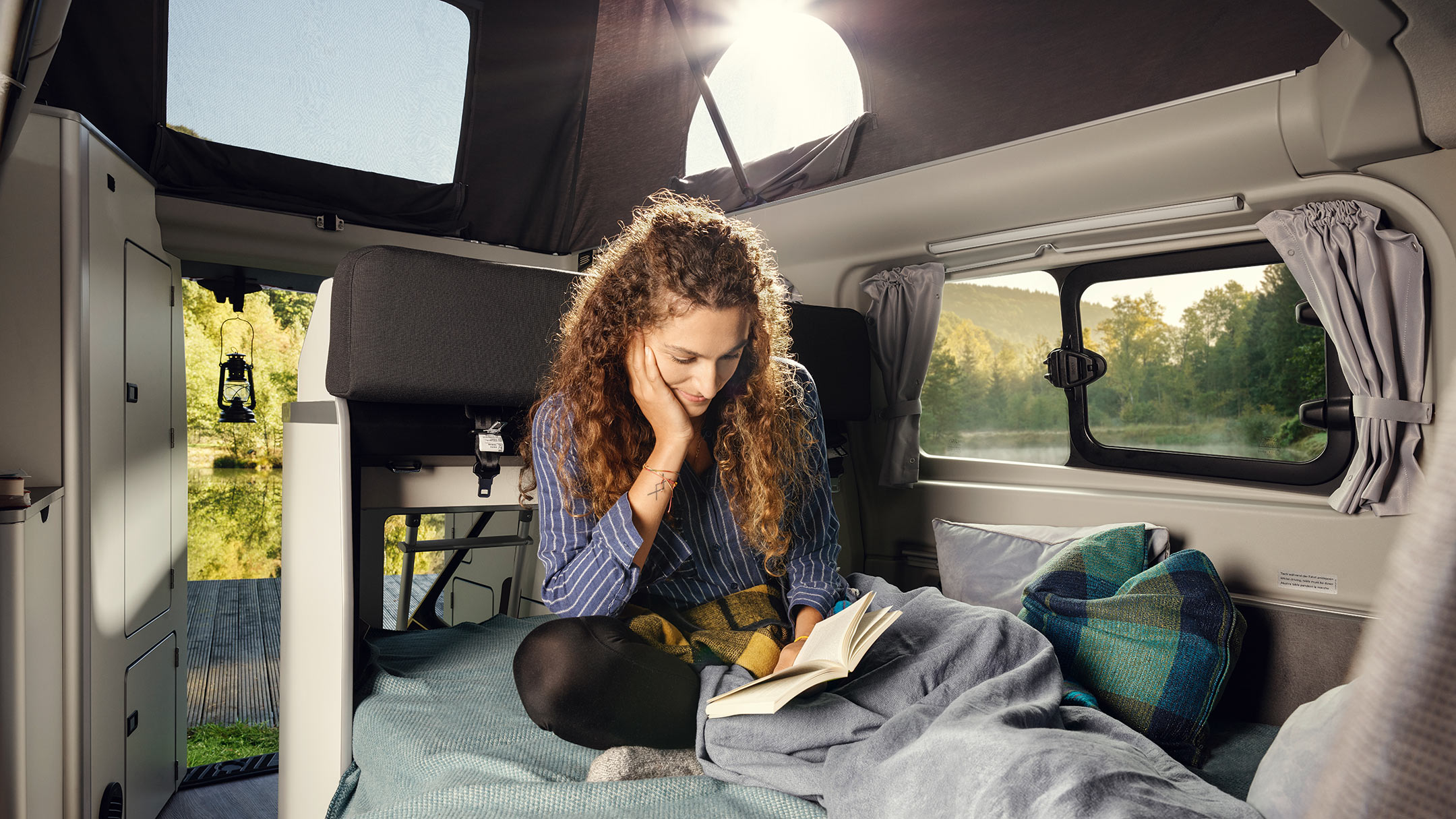 Ford Transit Custom Nugget Aufstelldach Innenraumansicht Frau sitzt auf Doppelbett in Wohnbereich Ford Transit Custom Nugget Aufstelldach Innenraumansicht Frau sitzt auf Doppelbett in Wohnbereich