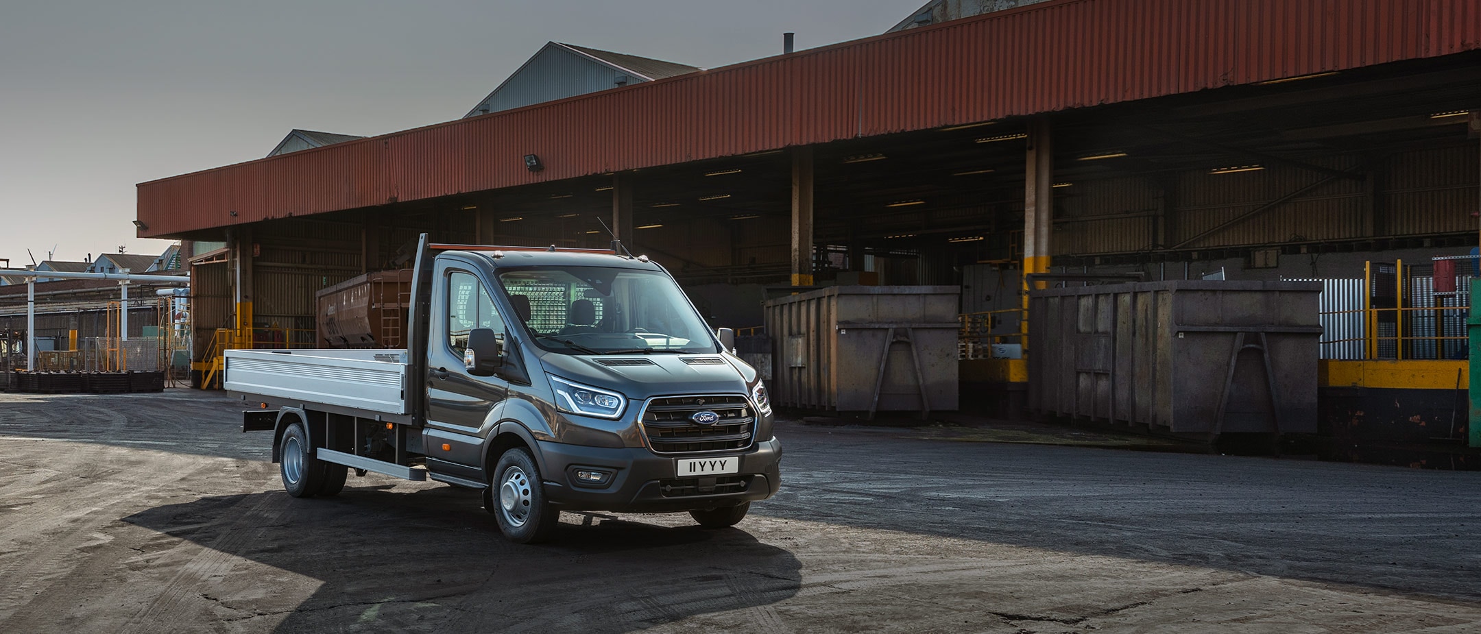 Ford Transit 5 Tonnen Pritschenwagen Schwarz ¾-Frontansicht parkt auf Betriebsgelände neben Überdachung mit Paletten