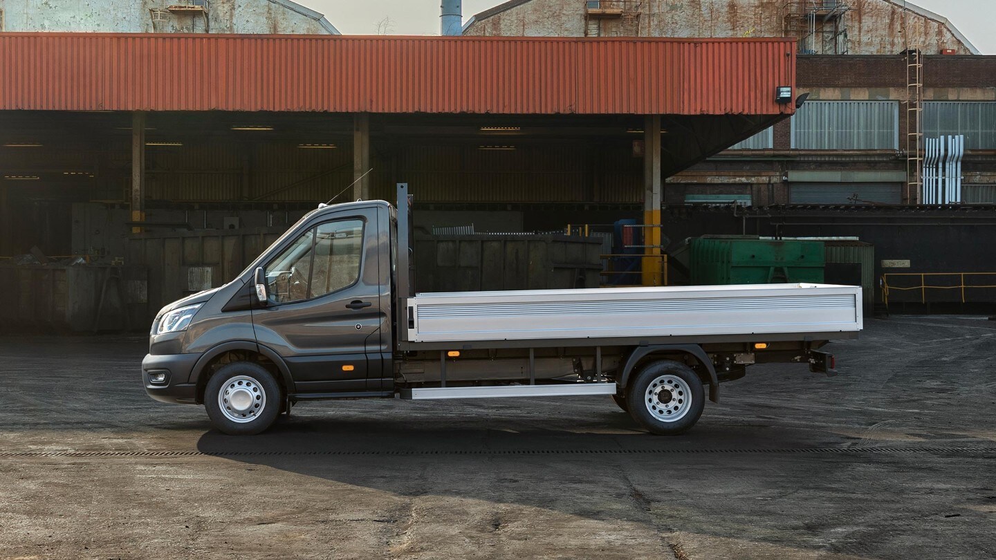 Ford Transit 5 Tonnen Pritschenwagen Schwarz Seitenansicht parkt auf Betriebsgelände neben Überdachung mit Paletten