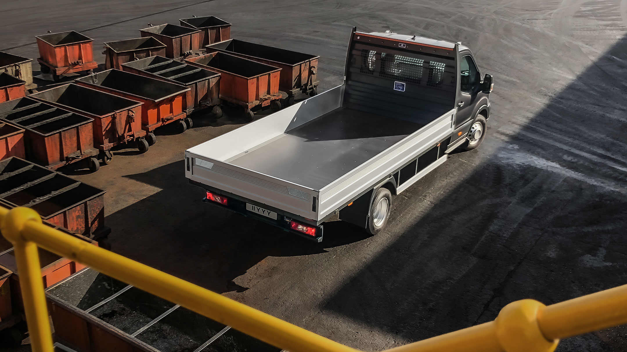 Ford Transit 5 Tonnen Pritschenwagen Schwarz ¾-Heckansicht Vogelperspektive parkt auf Betriebsgelände neben beladenen Paletten