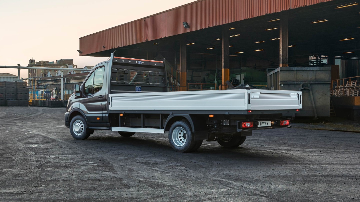 Ford Transit 5 Tonnen Pritschenwagen Schwarz ¾-Heckansicht parkt auf Betriebsgelände neben Überdachung mit Paletten