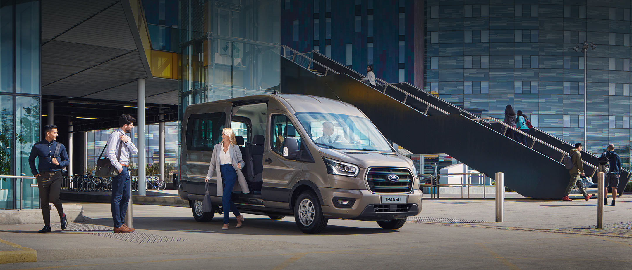 Ford Transit Bus Champagner ¾-Frontansicht geöffnete Schiebetür und Personen parkt vor modernem Gebäude