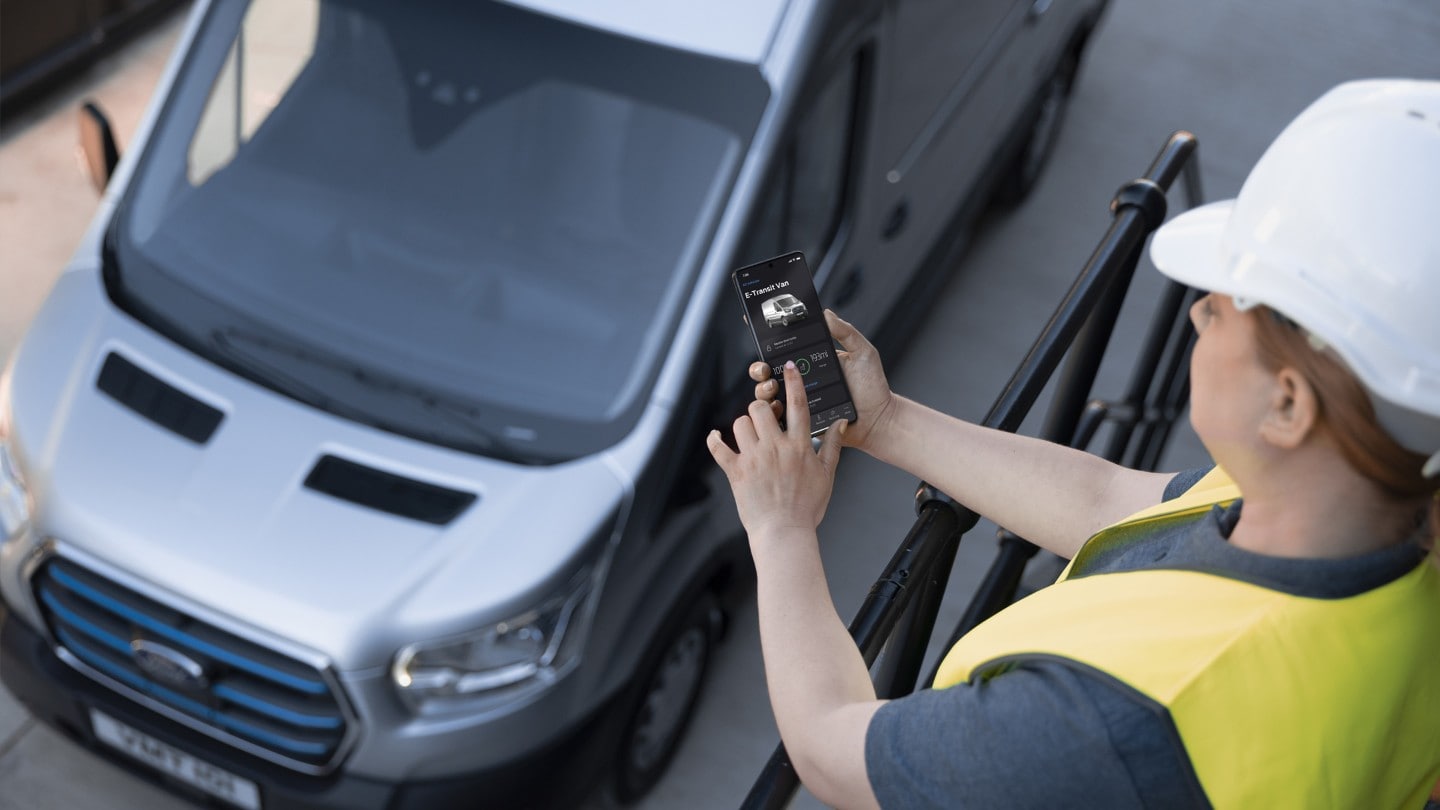 Frau mit Arbeitshelm steht neben Ford E-Transit und hält Mobiltelefon in der Hand