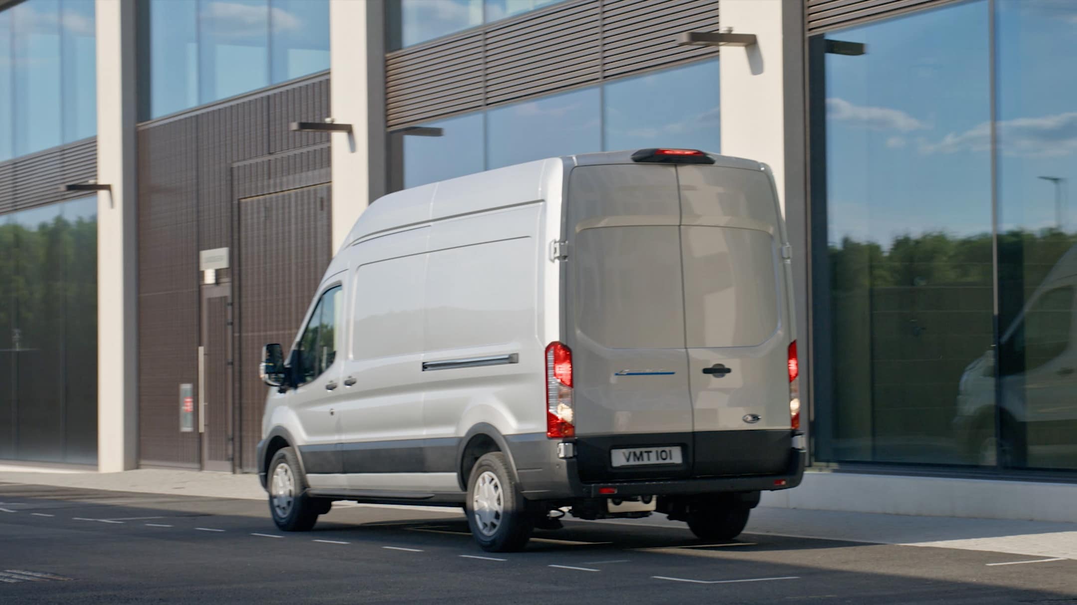 Ford E-Transit in Silber ¾-Heckansicht parkt vor modernem Gebäude