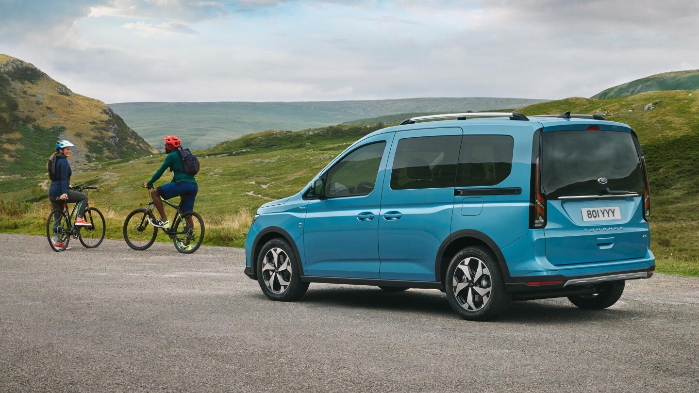 Ford Tourneo Connect in Hellblau ¾-Heckansicht steht auf Landstraße, davor stehen zwei Fahrradfahrer