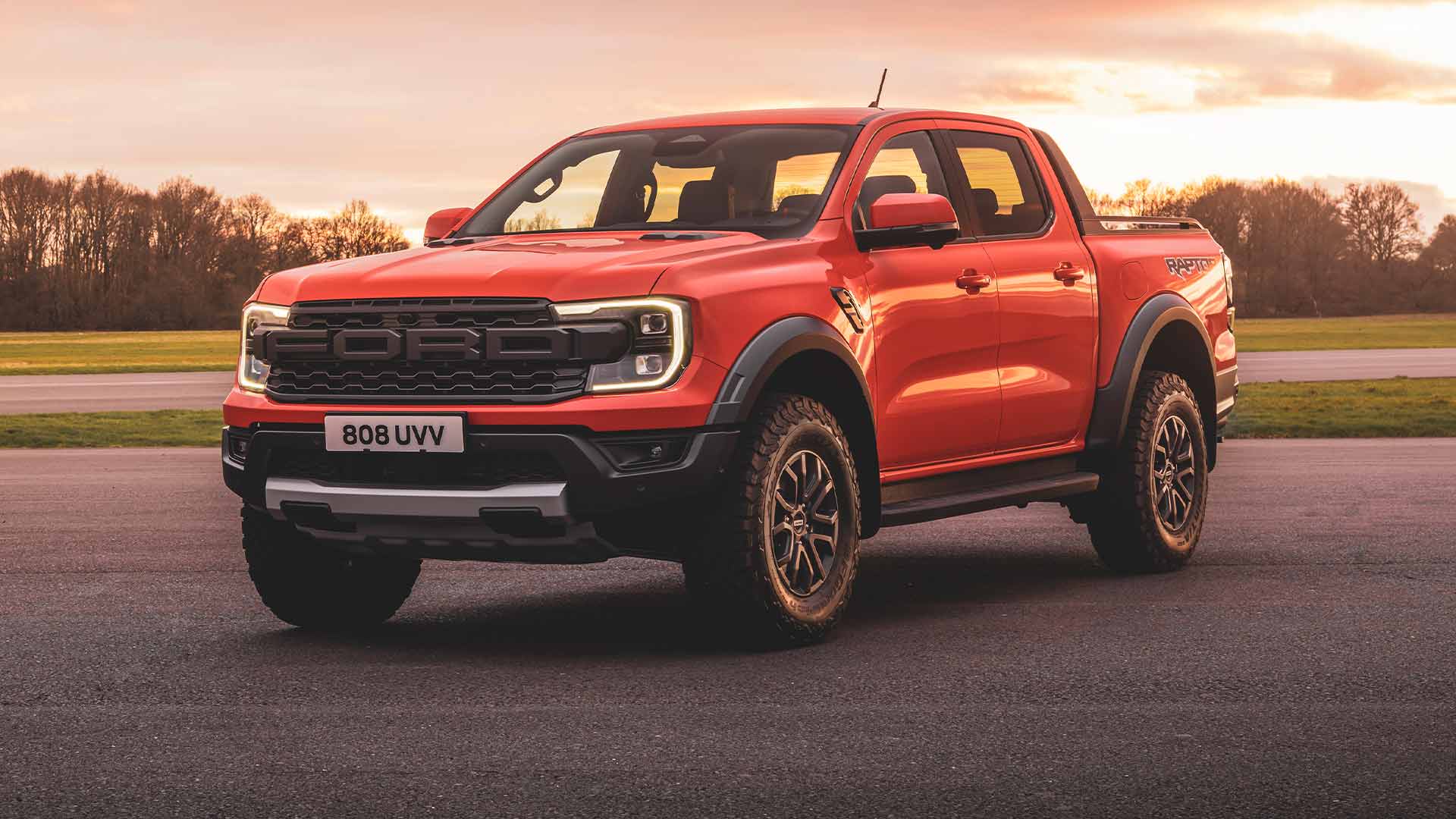 Code Orange Ford Ranger Raptor mit eingeschalteten Scheinwerfern auf einer Rennstrecke geparkt. 