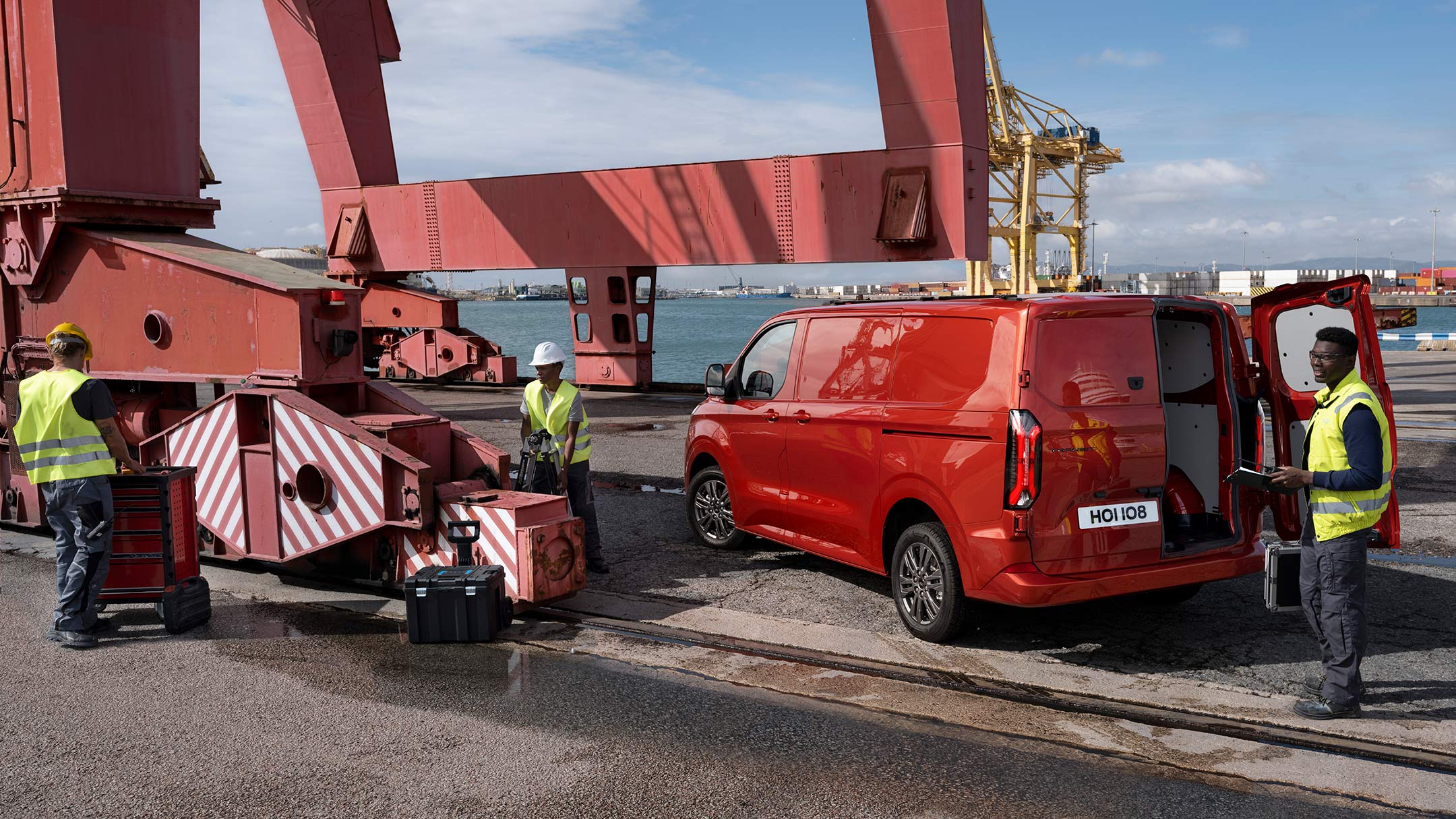 Ford Transit Custom in Rot in der ¾-Heckansicht wird von Hafenarbeitern beladen