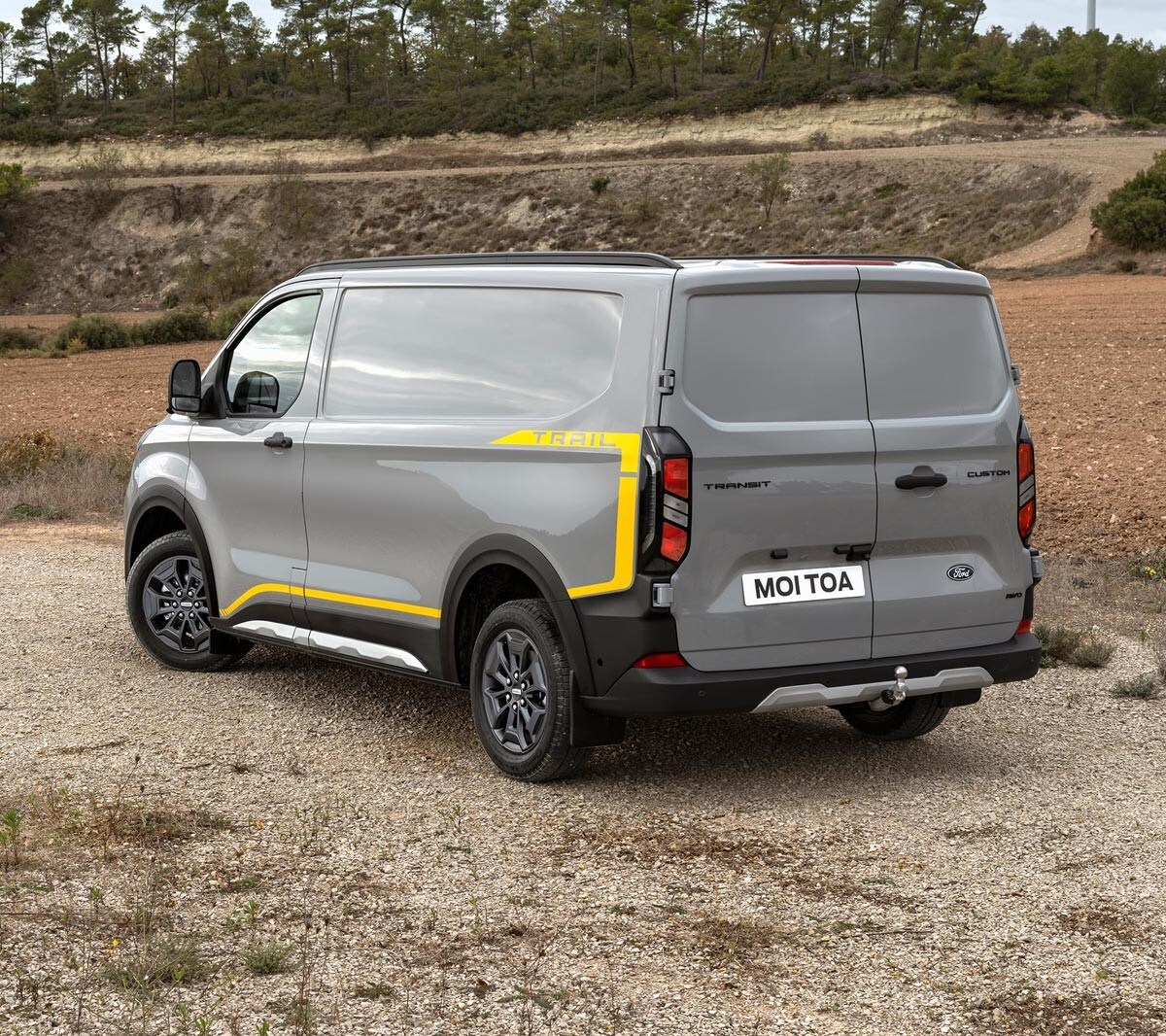 Ford Transit Custom in Grau in der ¾-Heckansicht parkt auf unbefestigtem Untergrund