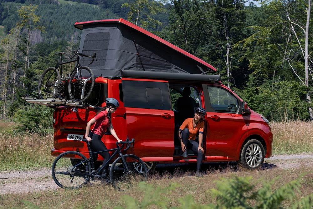 Personen laden Fahrräder vom Fahrradträger eines roten Ford Nugget ab.