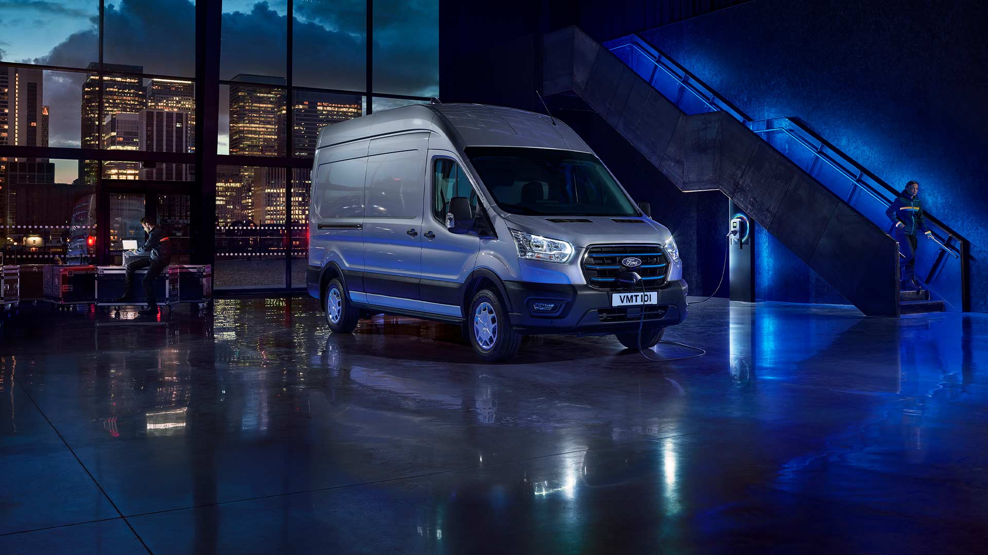 Ein silberner Ford E-Transit parkt in einer Halle; durch große Fenster ist die Skyline der Stadt zu sehen. 