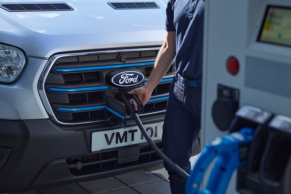 Nahaufnahme einer Person, die ein Ladekabel an einen silbernen Ford E-Transit  Kastenwagen anschließt.