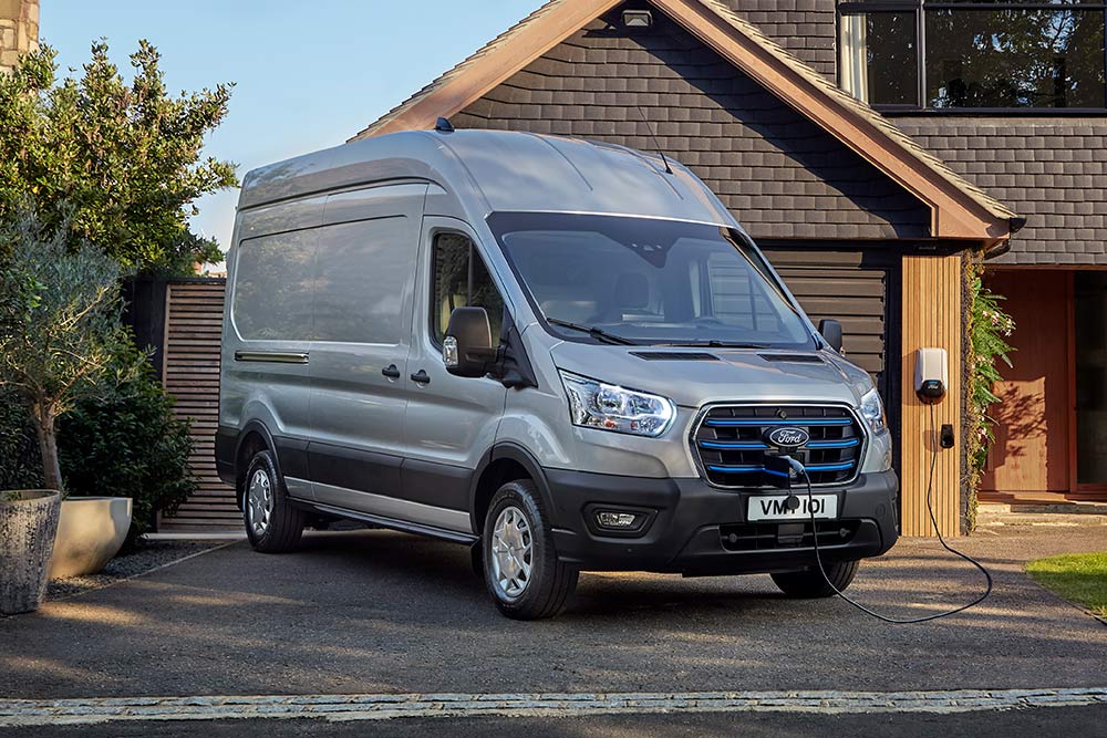Ein silberner Ford E-Transit parkt in einer Einfahrt neben einem Haus und ist an eine Ladestation angeschlossen.