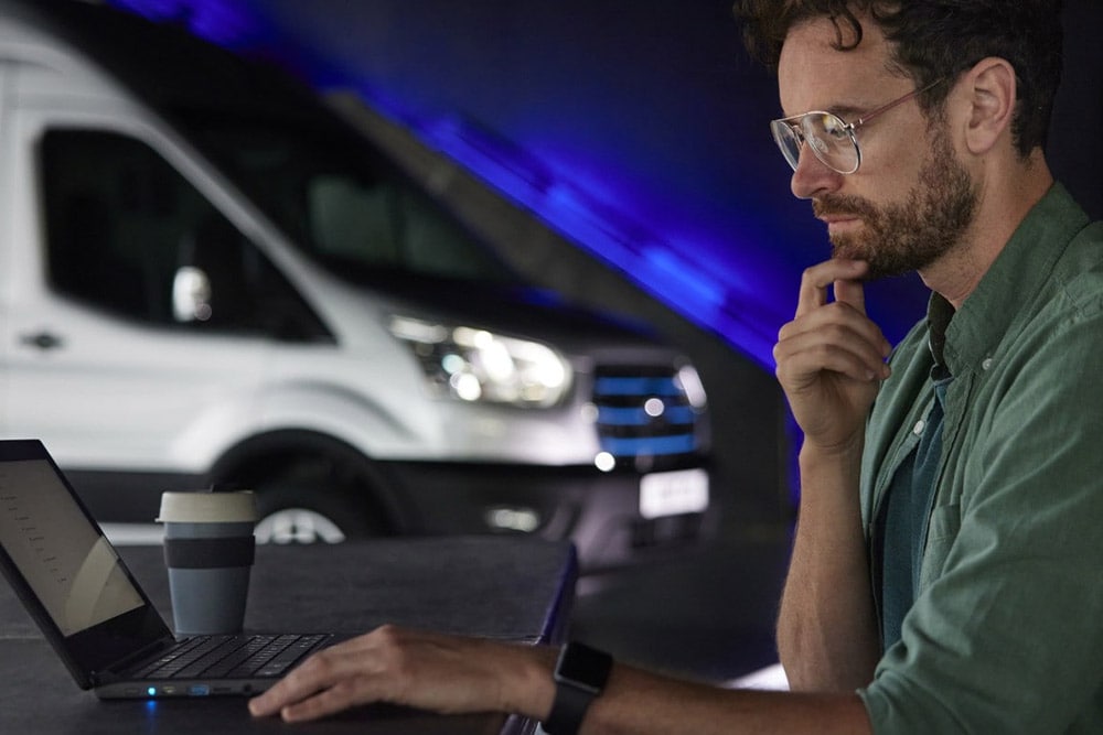 Ein Mann arbeitet an einem Laptop mit einem Ford E-Transit Kastenwagen im Hintergrund.