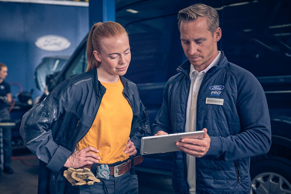 Zwei Mechaniker in einer Werkstatt schauen vor einem Ford E-Transit Kastenwagen auf ein Tablet.