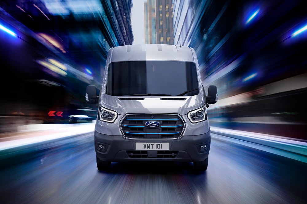 Frontansicht eines grauen Ford Transit-Transporters mit blauen Akzenten am Kühlergrill, der nachts mit hoher Geschwindigkeit durch eine Stadt fährt.