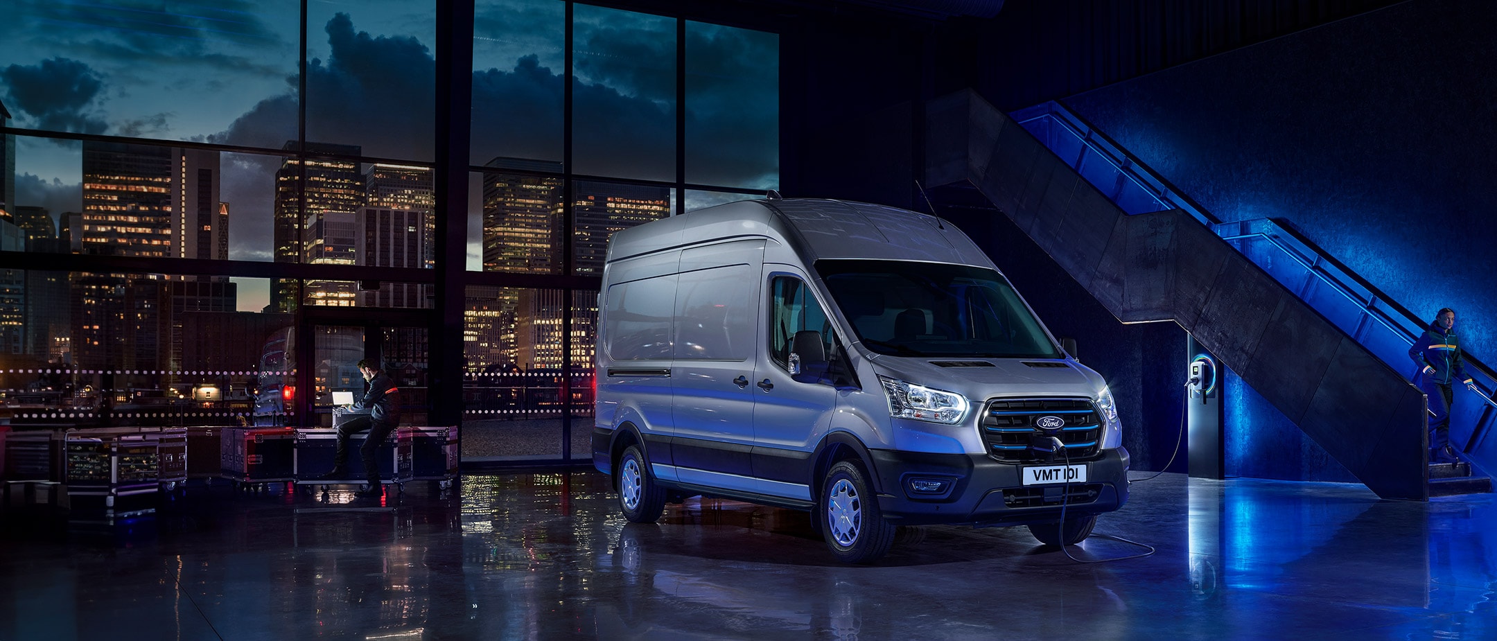 Ein silberner Ford E-Transit wird in Innenräumen aufgeladen, mit einer Person, die an einem Laptop arbeitet, und einer Skyline im Hintergrund. Text: Sorgenfreies Fahren.
