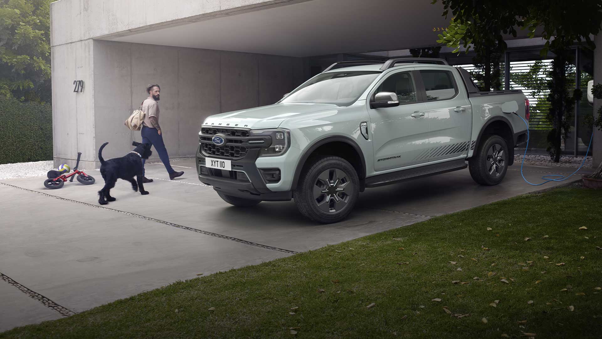 Ein Mann und ein Hund gehen zu einem grauen Ford Ranger Plug-in-Hybrid-Pick-up-Truck, der in einer Einfahrt aufgeladen wird. 