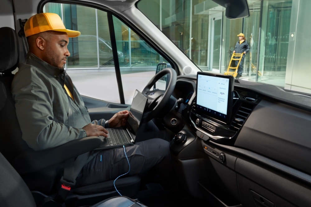 Ein Mann sitzt auf dem Fahrersitz eines Ford Transit Kastenwagens und arbeitet an einem Laptop.